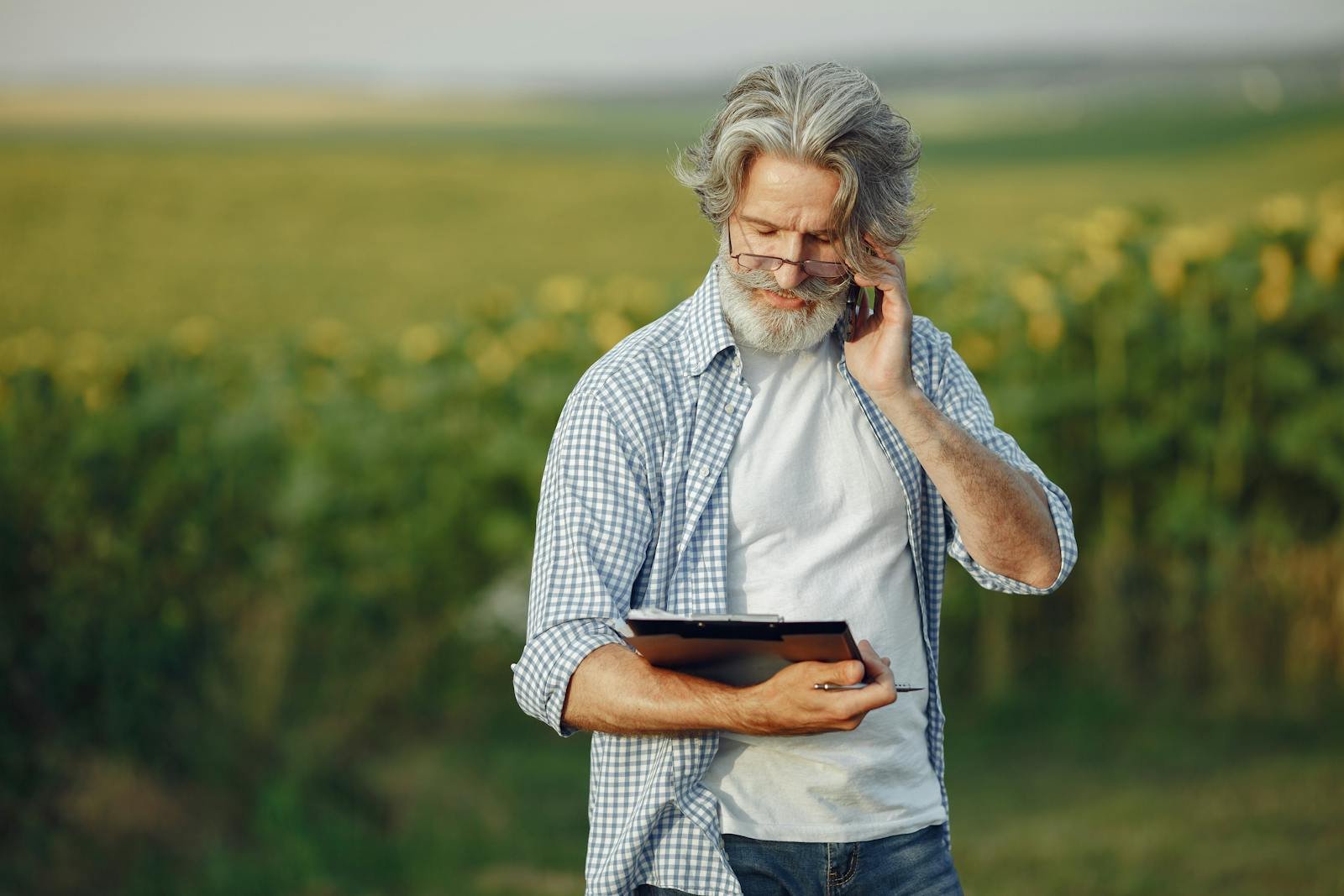 Elderly man in a field using a smartphone and tablet, wearing glasses and a casual shirt.